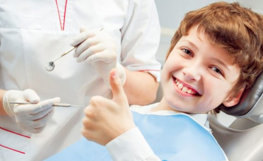 A happy kid showing his thumbs, up sitting on a chair near a dentist who is having some dental tools at a dental clinic.