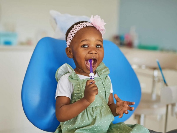 A happy kid sitting on a colorful blue chair, keeping a brush in its mouth.